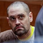 Man with shorn head and stubble in T-shirt with neon yellow collar looks upward at someone in a courtroom.