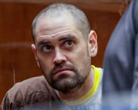 Man with shorn head and stubble in T-shirt with neon yellow collar looks upward at someone in a courtroom.