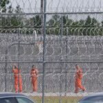 Men in orange jumpsuits behind a high chain-link fence laced through with razor wire.