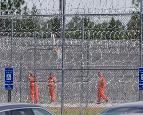 Men in orange jumpsuits behind a high chain-link fence laced through with razor wire.