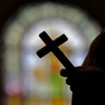Stock image of silhouetted hand holding cross, with stained glass blurry in background.