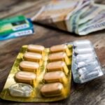 Pill blister packs sit on a wooden table beside banknotes and credit cards