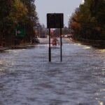 The A82 at Dumbuck Quarry is flooded, with water across the entire road. In the distance, there are a few cars on the road ahead.