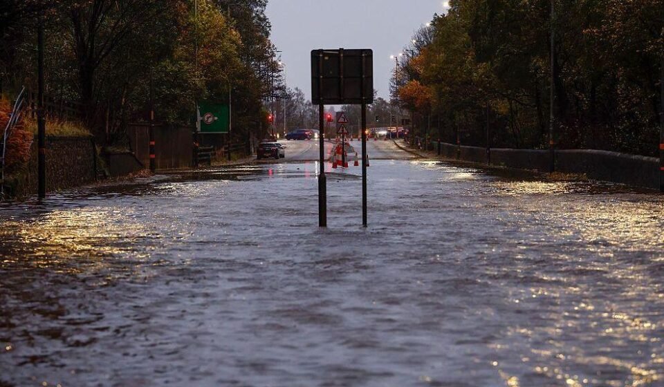 The A82 at Dumbuck Quarry is flooded, with water across the entire road. In the distance, there are a few cars on the road ahead.